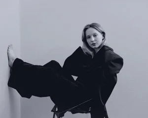 Monochrome portrait of a woman seated in a chair with one bare foot propped against the corner wall, wearing an oversized dark blazer.
