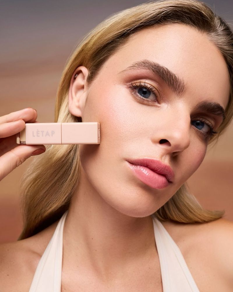 Close-up beauty portrait of a blonde woman holding a pale pink Letap cosmetic stick beside her cheek against a warm gradient backdrop.
