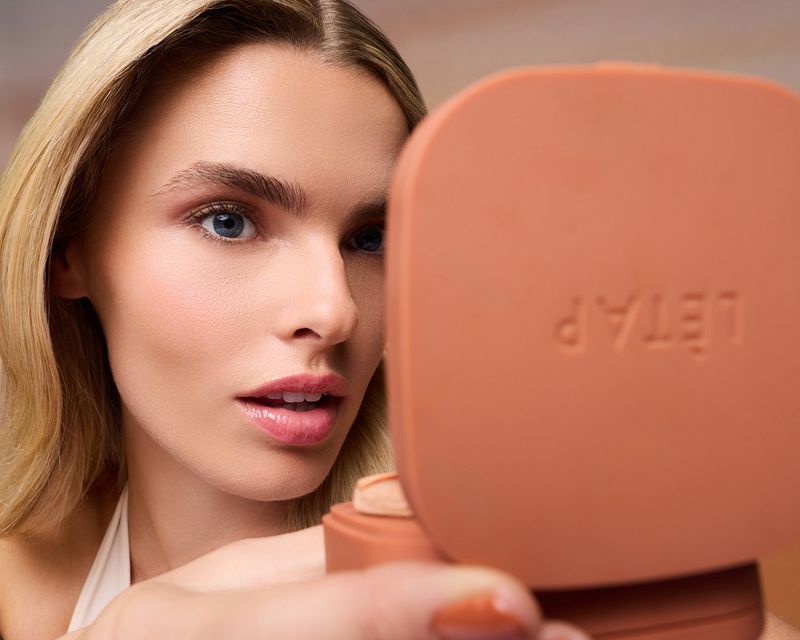 Close-up beauty portrait of a blonde woman with blue eyes looking into an open peach makeup compact mirror.
