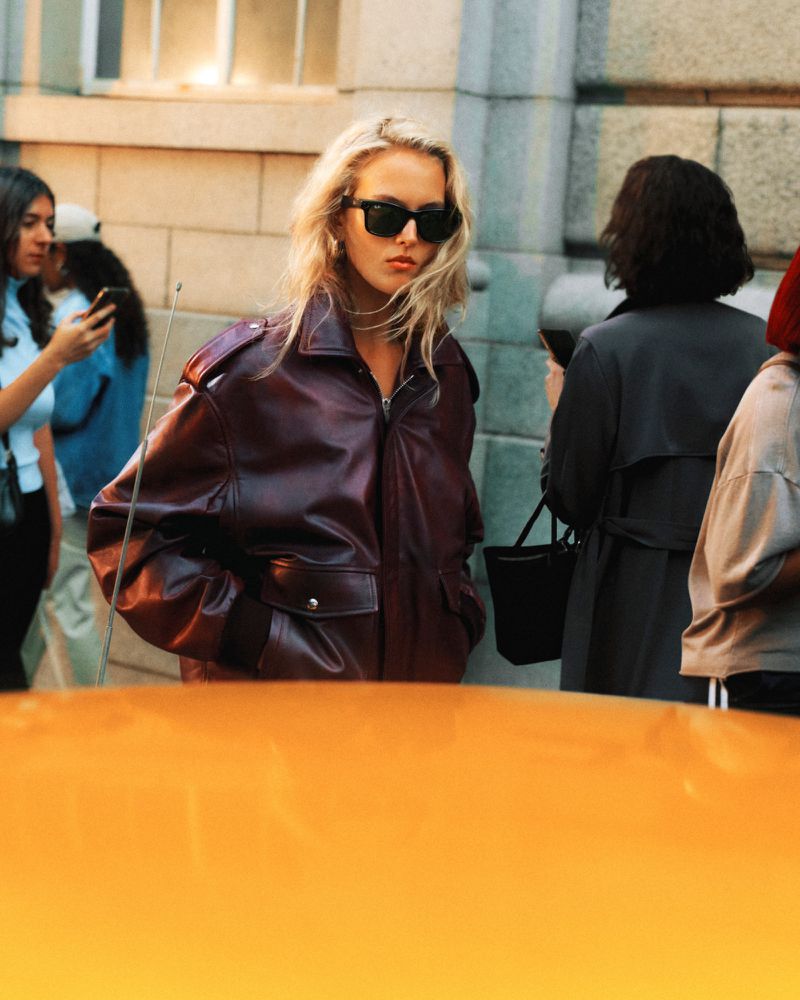 Blonde woman wearing black sunglasses and an oversized burgundy leather bomber jacket on a city street behind an orange car.