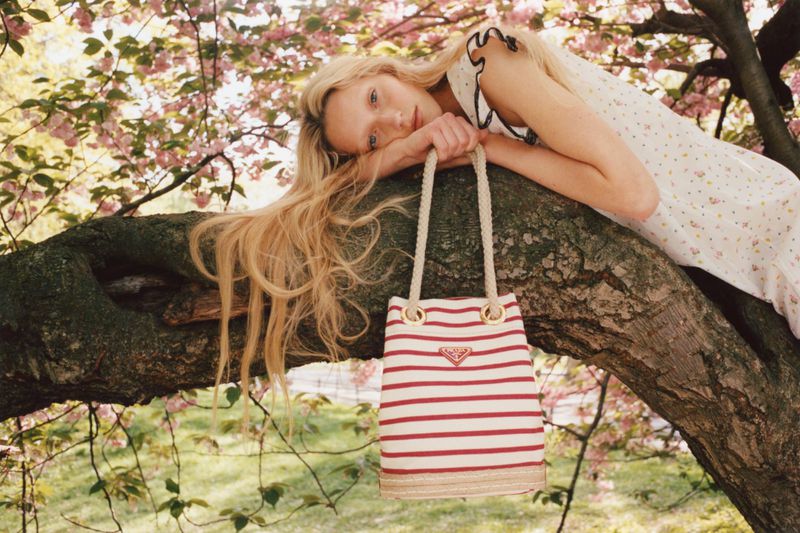 Young woman with long blonde hair lies on a tree branch with pink blossoms, holding a red-and-white Prada striped tote bag outdoors.