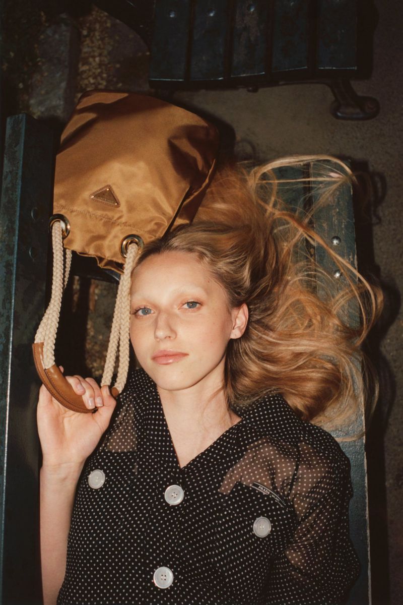 Portrait of a fair-skinned person with long blonde hair, a black polka dot dress, and a beige purse against a bench-like background.