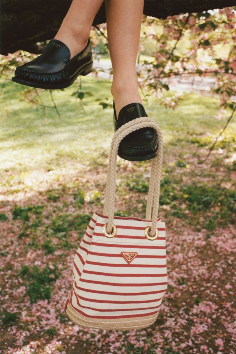 Black leather loafers on feet and a striped canvas bucket bag hanging from a tree branch in an outdoor fashion shot.