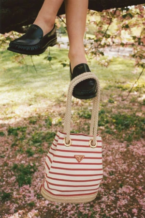 Black leather loafers on feet and a striped canvas bucket bag hanging from a tree branch in an outdoor fashion shot.