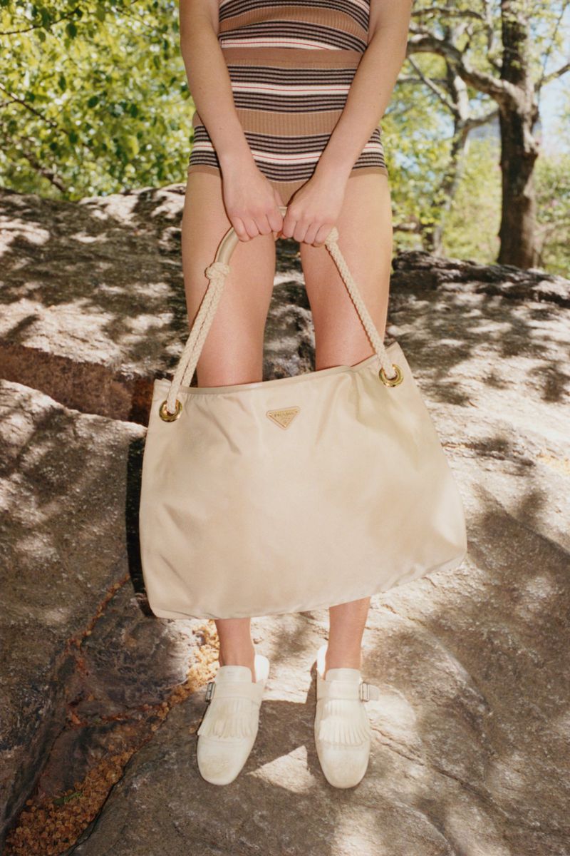 A woman standing on a rock formation holds a Prada handbag with a beige and brown striped swimsuit and white shoes in natural light.