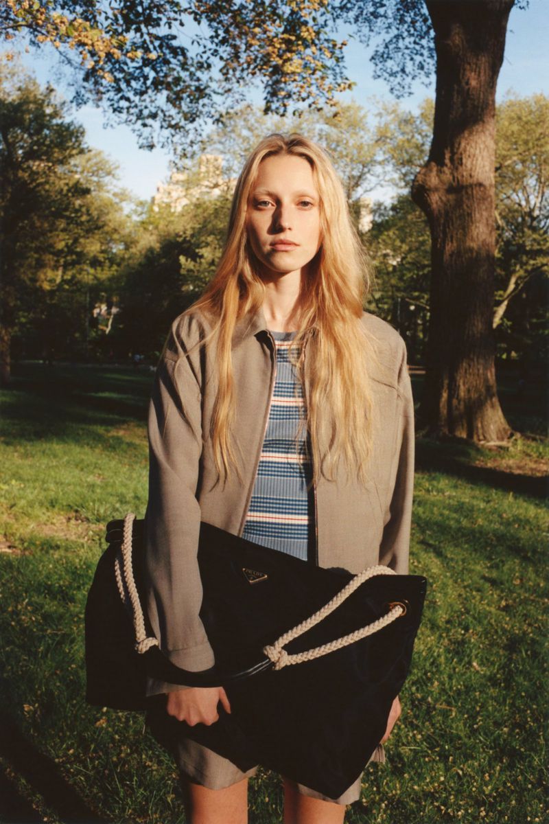 Young woman in an outdoor park setting wearing a grey jacket and striped shirt, carrying a black Prada tote bag with rope straps.