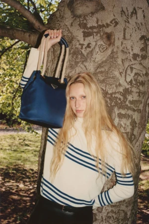 A woman in a striped sweater holds a blue Prada bag against a tree in a park setting with natural light, showcasing fashion and nature.
