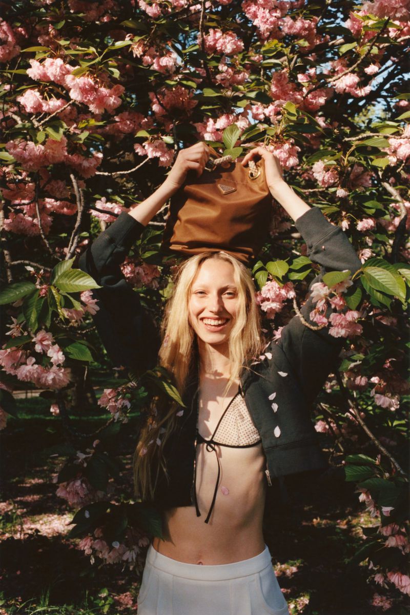 A young adult woman smiles while posing outdoors with a brown Prada handbag balanced on her head, framed by pink cherry blossoms.