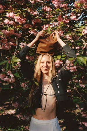 A young adult woman smiles while posing outdoors with a brown Prada handbag balanced on her head, framed by pink cherry blossoms.