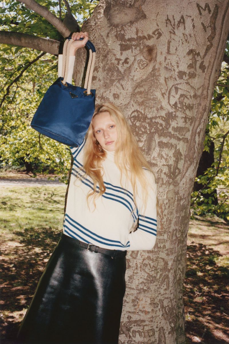 A fair-skinned woman with blonde hair poses with a blue tote bag near a tree in a sunlit outdoor setting.