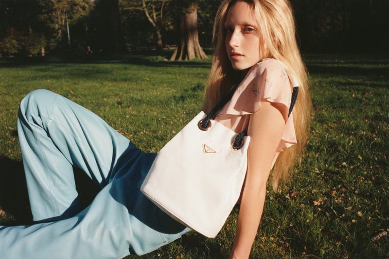 Woman in floral top and teal pants sits on grass with white Prada handbag in outdoor setting, illuminated by soft, diffused sunlight.