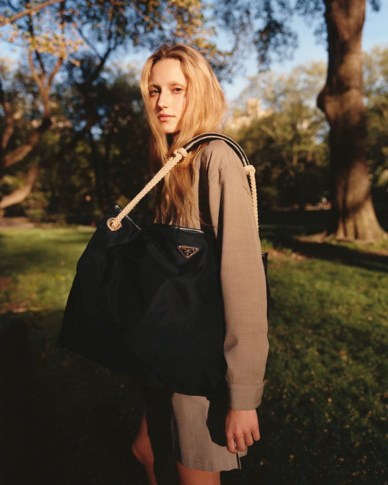A blonde woman carrying a Prada handbag stands outdoors in a park with trees and greenery in the background, illuminated by natural light.