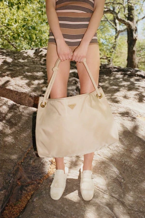 Model standing outdoors on rocks holding a large beige shoulder bag wearing a striped top and white loafers