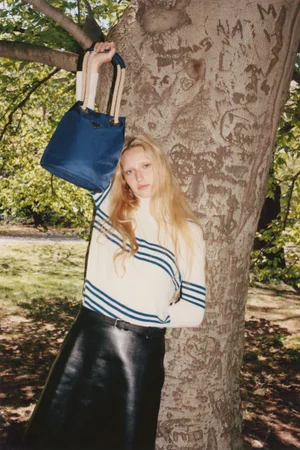 A blond woman wearing a striped turtleneck and black skirt poses against a large tree holding up a blue handbag.