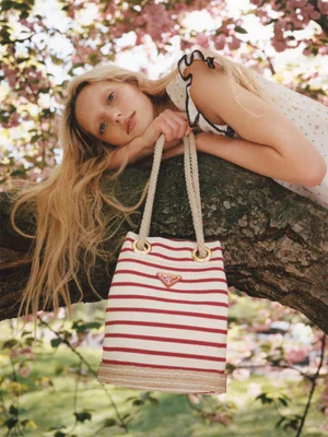 Blonde woman reclining on a tree branch holding a striped canvas bucket bag against a background of pink blossoms
