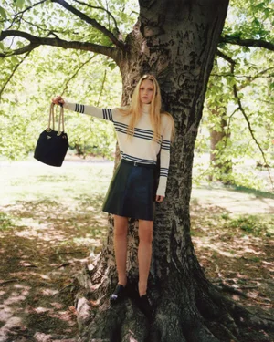 Model standing against a large tree holding a black handbag in a park setting for Elle UK