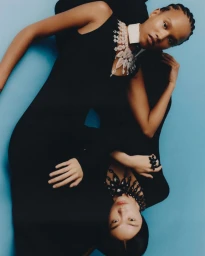 Two models lying inverted against a blue backdrop wearing black sleeveless tops, statement crystal and beaded necklaces, and rings.