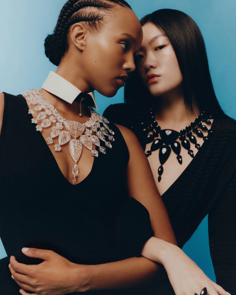 Two fashion models posing in black dresses and ornate statement necklaces against a vibrant solid blue studio background.