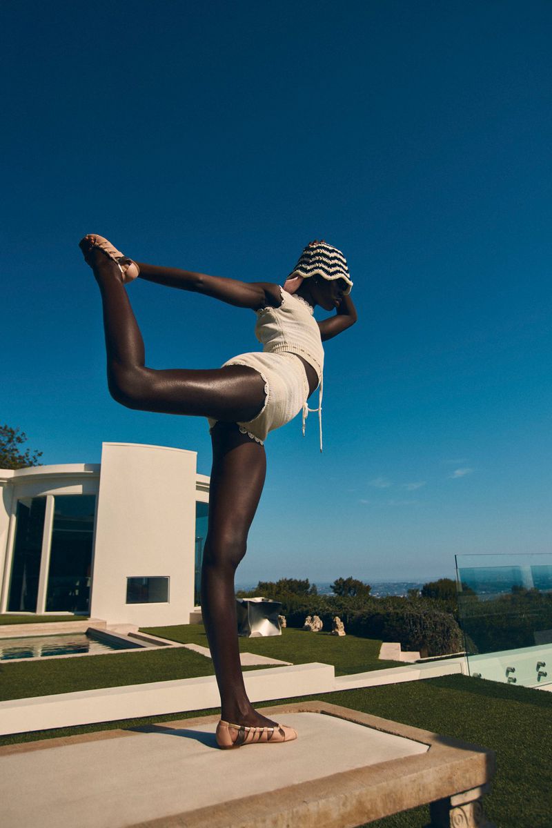 A person with dark skin performing a dancer pose in a white knit set and patterned bucket hat against a clear deep blue sky