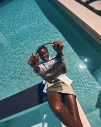 Dija Kallon lies poolside holding up a brown leather handbag toward the camera above sparkling turquoise water.
