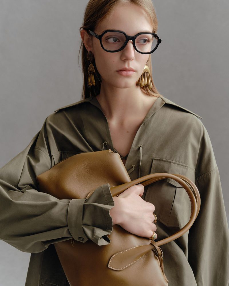 Model wearing black glasses and an olive green shirt holding a tan leather tote bag against a grey background