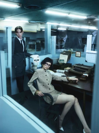 Two women pose in a retro-style office setting. One sits at a desk with a typewriter, while the other stands beside a coffee maker.