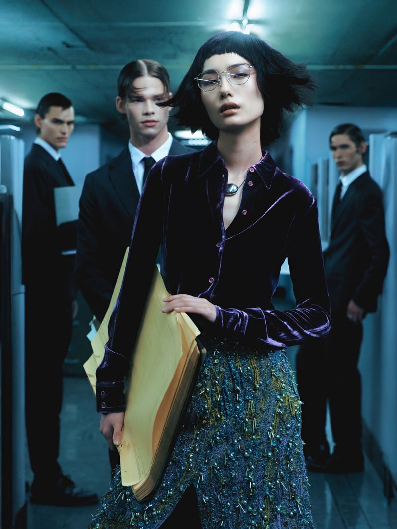 A fashionable woman with short dark hair holds files in an office hallway, flanked by three men in suits for a fashion editorial.