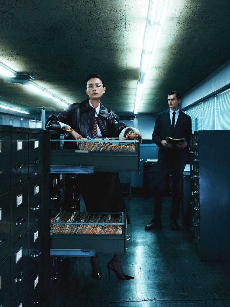 Stylish woman in a leather jacket leans on filing cabinet, while a man in a suit stands in the background in a vintage office setting.