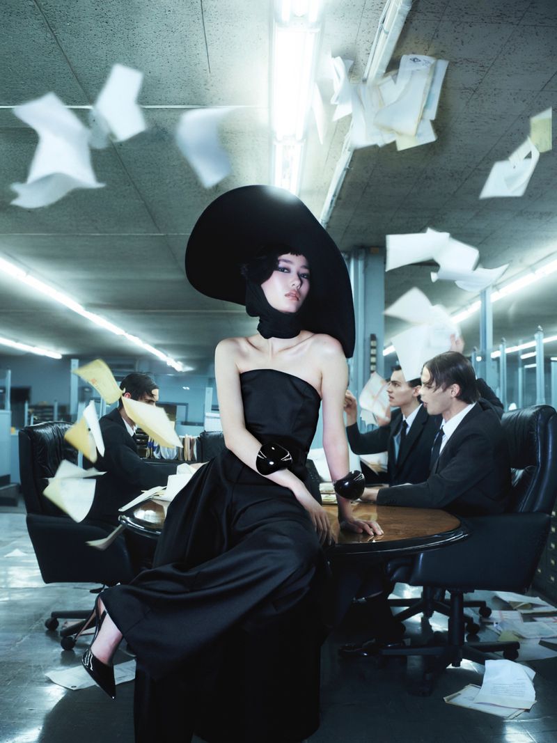 Elegant woman in a black dress and hat sits on a table surrounded by suited men in a modern office filled with floating papers.
