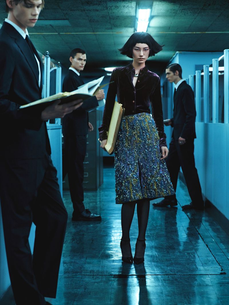 Fashion editorial shot in a hallway with a woman in a patterned skirt and several men in suits holding documents, under fluorescent lights.