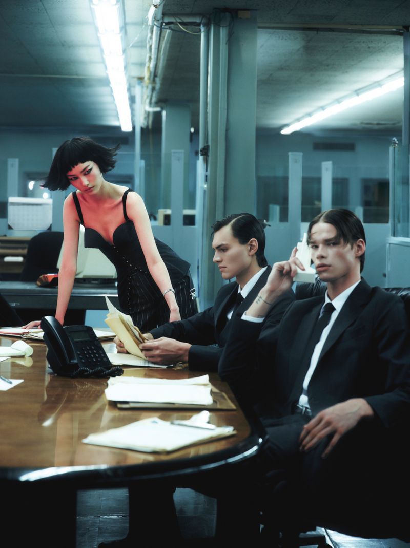Fashion shoot of a young woman in a black dress and two men in suits at a conference table with paperwork and a black landline phone.