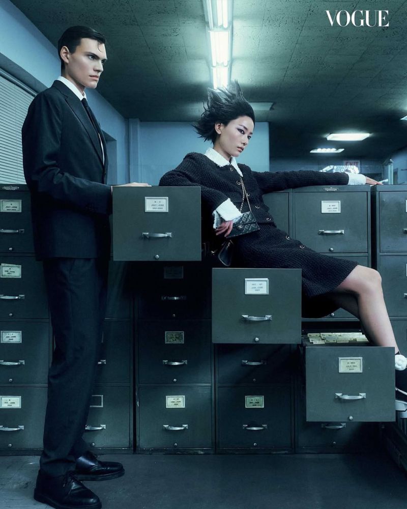 Editorial shot of a male model in a suit standing near filing cabinets and a female model sitting on the cabinets indoors.