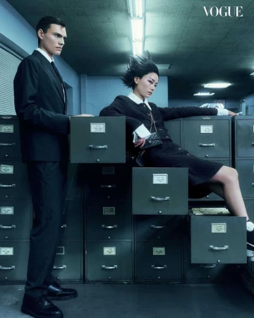 Editorial shot of a male model in a suit standing near filing cabinets and a female model sitting on the cabinets indoors.