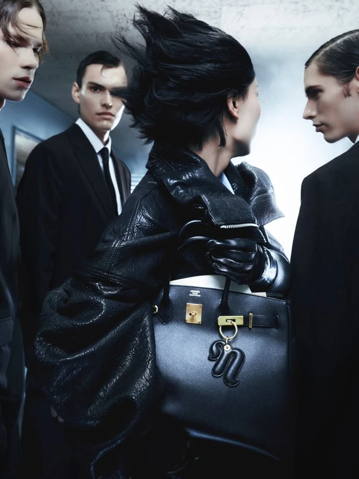 Model in black leather jacket holding a black leather handbag surrounded by three men in dark suits
