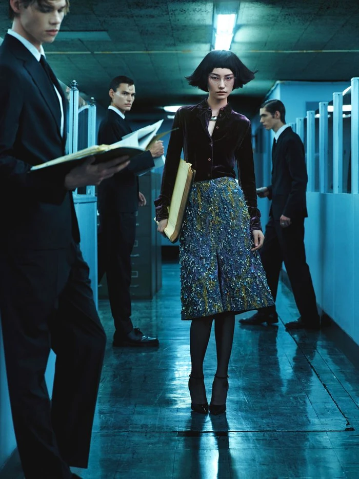 A woman with short dark hair in a velvet top and patterned skirt stands in an office corridor among men in suits holding folders.