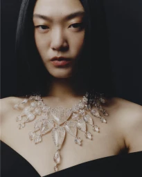 East Asian model wearing an elaborate clear crystal statement necklace and black off-shoulder top against a dark backdrop.