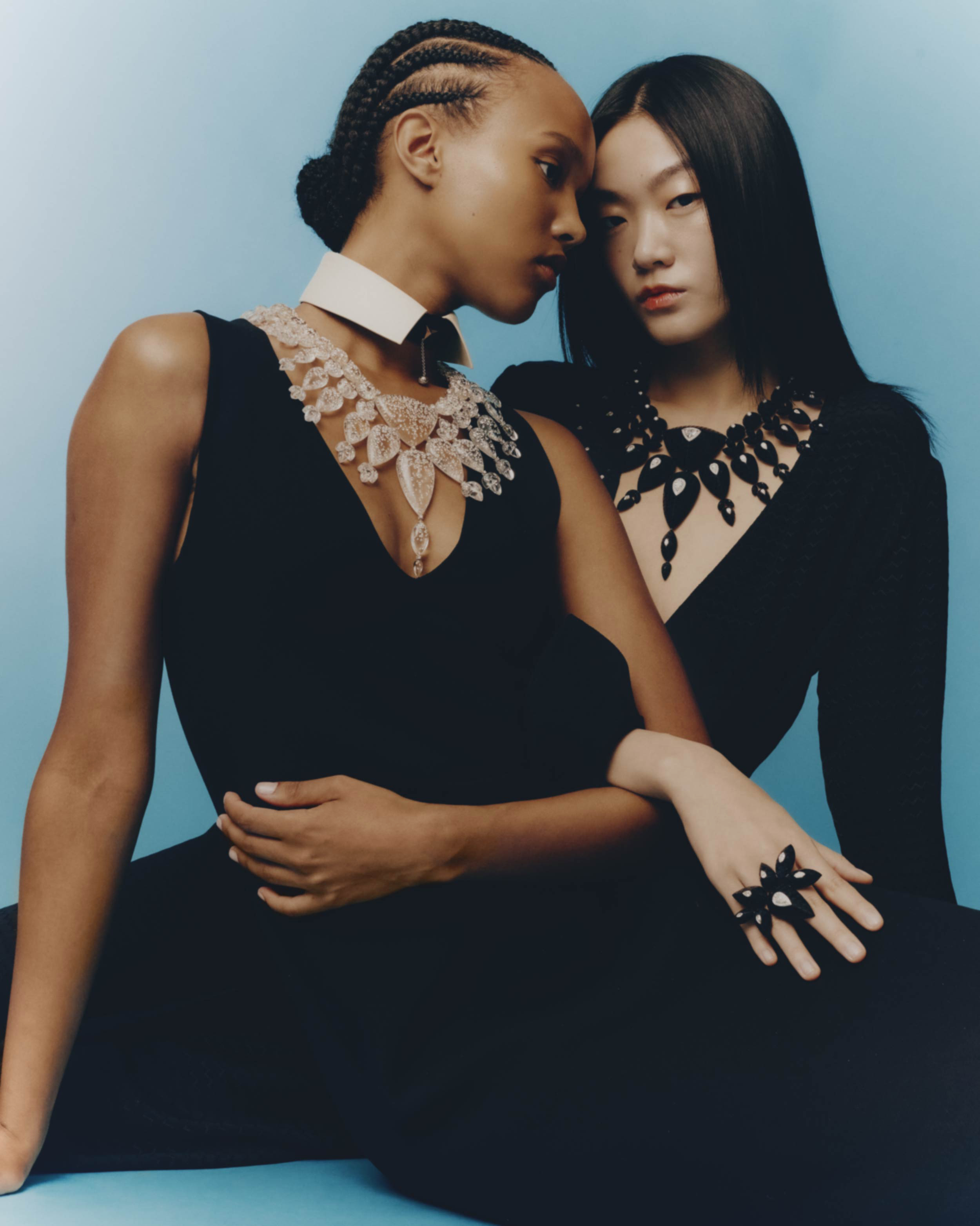 Two female models in black dresses wearing ornate statement necklaces against a vibrant blue studio background.