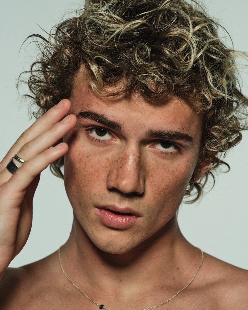 Close-up portrait of a shirtless young man with curly highlighted hair and freckles raising a hand to his face against a pale studio backdrop.
