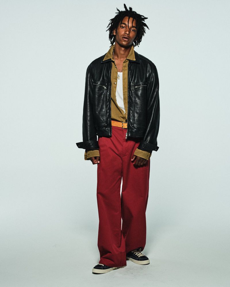 Young Black man with dreadlocks wearing a black leather jacket and wide-leg red pants against a pale studio seamless.