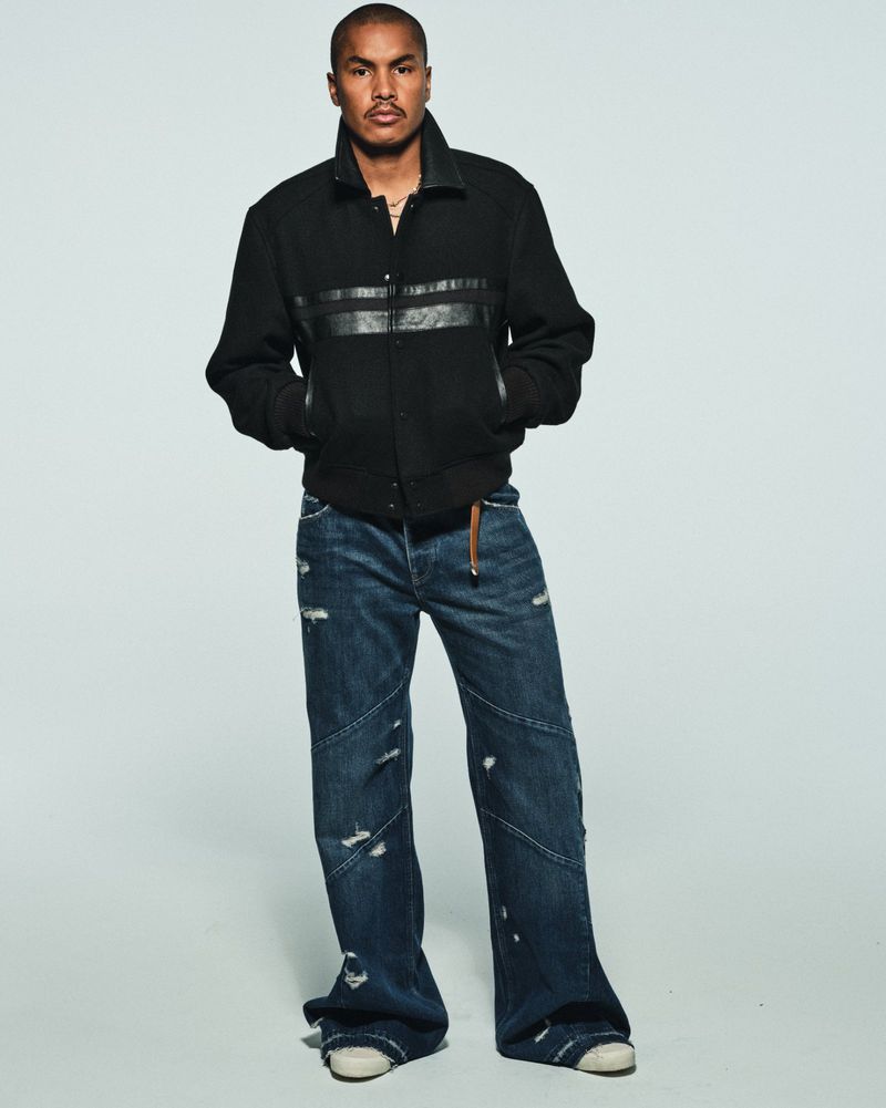 Man posing in a black jacket and wide-leg distressed denim jeans against a pale studio seamless backdrop.