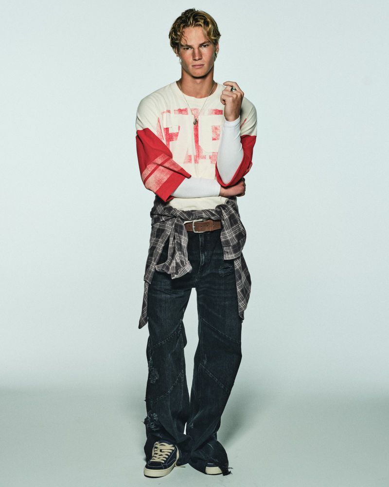 A young man wearing a red and white graphic top, plaid shirt tied around the waist, and baggy dark jeans against a studio backdrop.