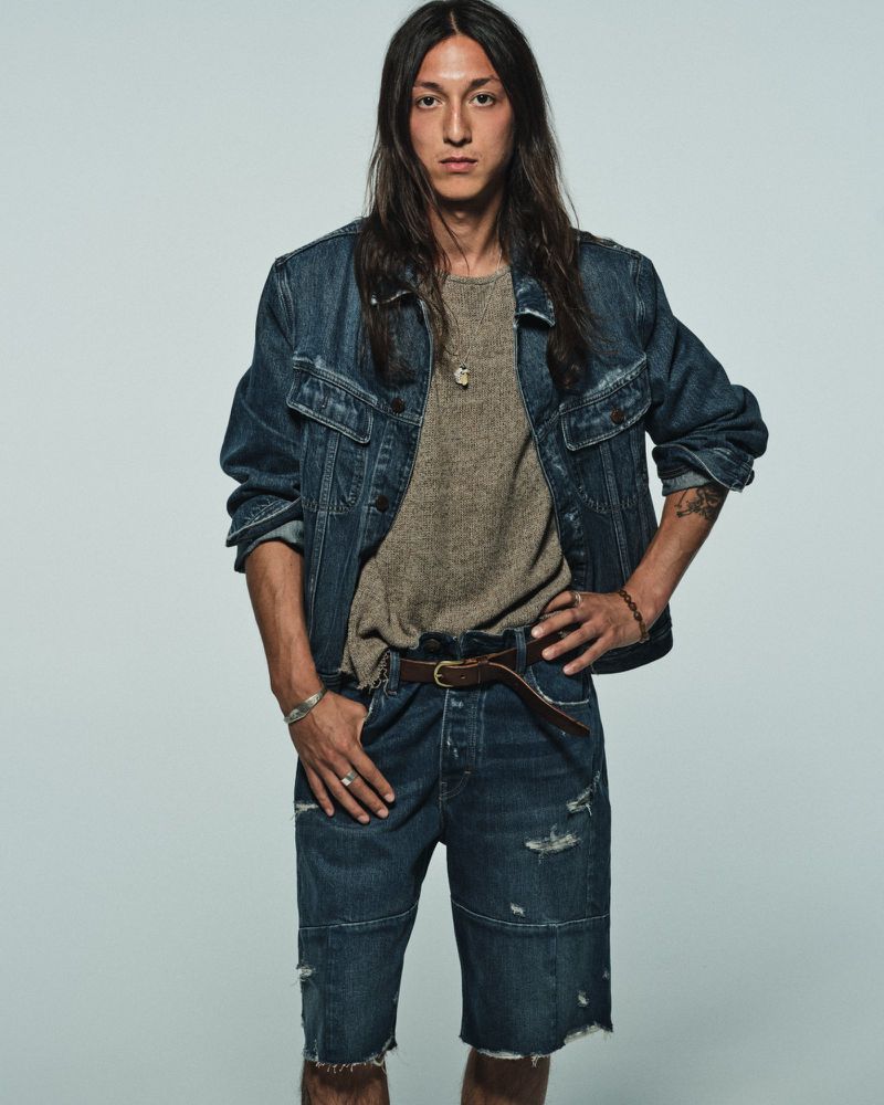 A young man with long dark hair wears a denim jacket and distressed denim shorts, standing against a plain light gray studio background.