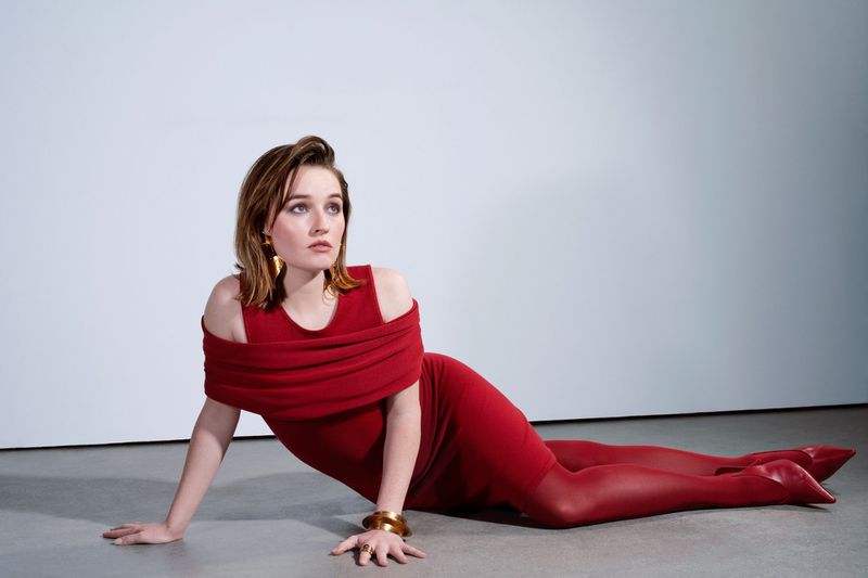 A woman in a red dress and matching tights is lying on the floor in a studio setting, with a solid white background and bright lighting.