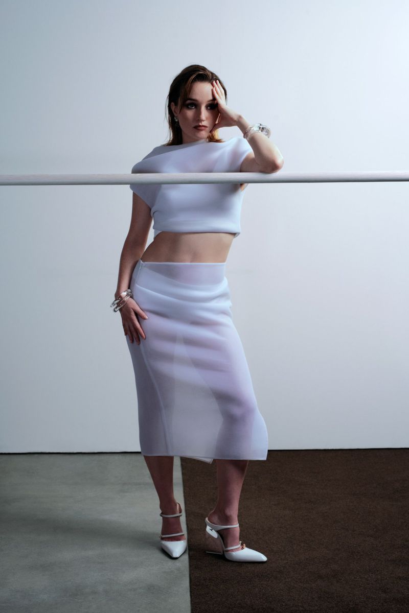 A woman poses indoors wearing a light blue two-piece outfit with a sheer skirt and white heels, leaning against a horizontal pole.