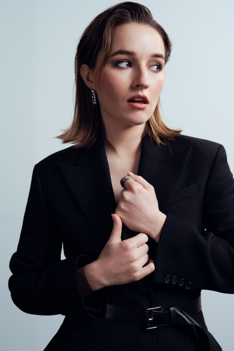 A young adult woman with short auburn hair poses in a black suit, gazing away in a studio shot against a light blue background.