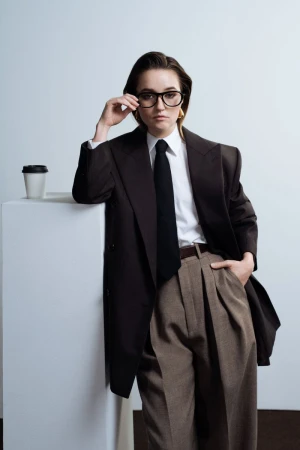 A woman in a dark business suit leans against a white block with a coffee cup, wearing glasses and posing against a pale background.