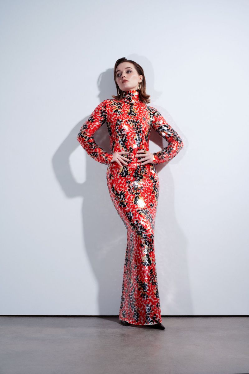 Elegant woman in a long red and black floral gown, posing in a studio against a white wall with hands on her hips.