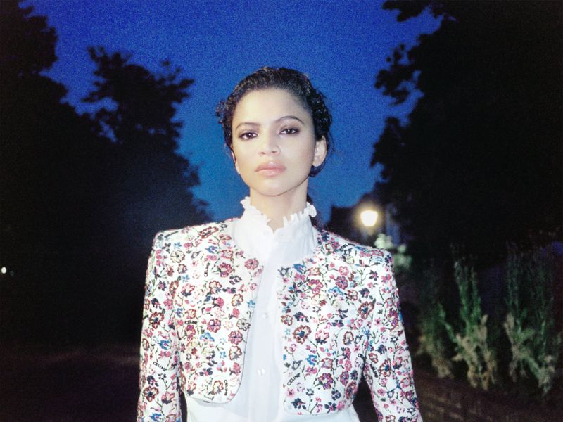 Model Bruna Souza in a floral cropped jacket and white ruffle shirt outdoors against a dark night sky.
