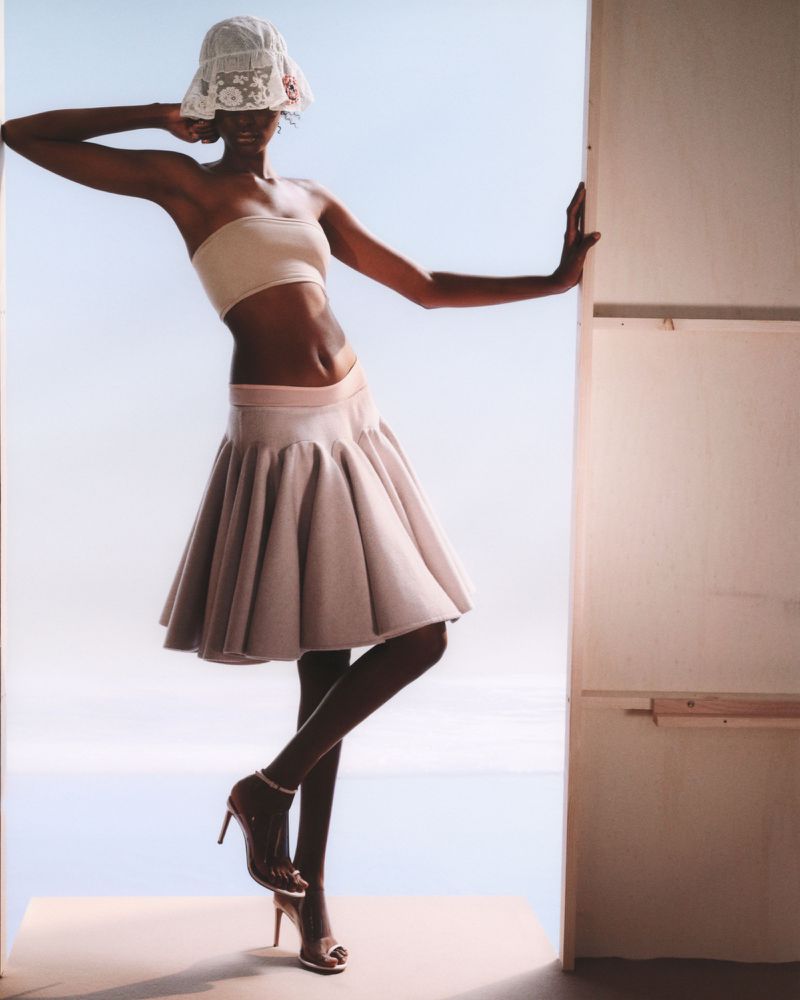 A woman in a beige top and skirt poses in front of a sky-blue backdrop with an open doorway, wearing a floral hat and high heels.