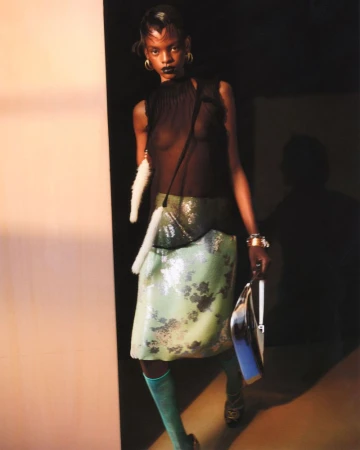 Elegant Black model strikes a pose in a sheer black top, shiny mint green skirt, carrying a silver bag, and wearing mint socks.